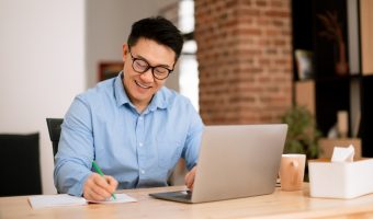 Happy asian man making notes, analyzing data for investment at workplace with laptop computer in